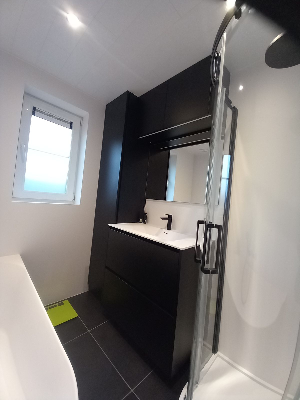 Badkamer met wanden in wit volkern zonder voegen, zwart badmeubel, hoge kast en ronde douchecabine in Langemark-Poelkapelle