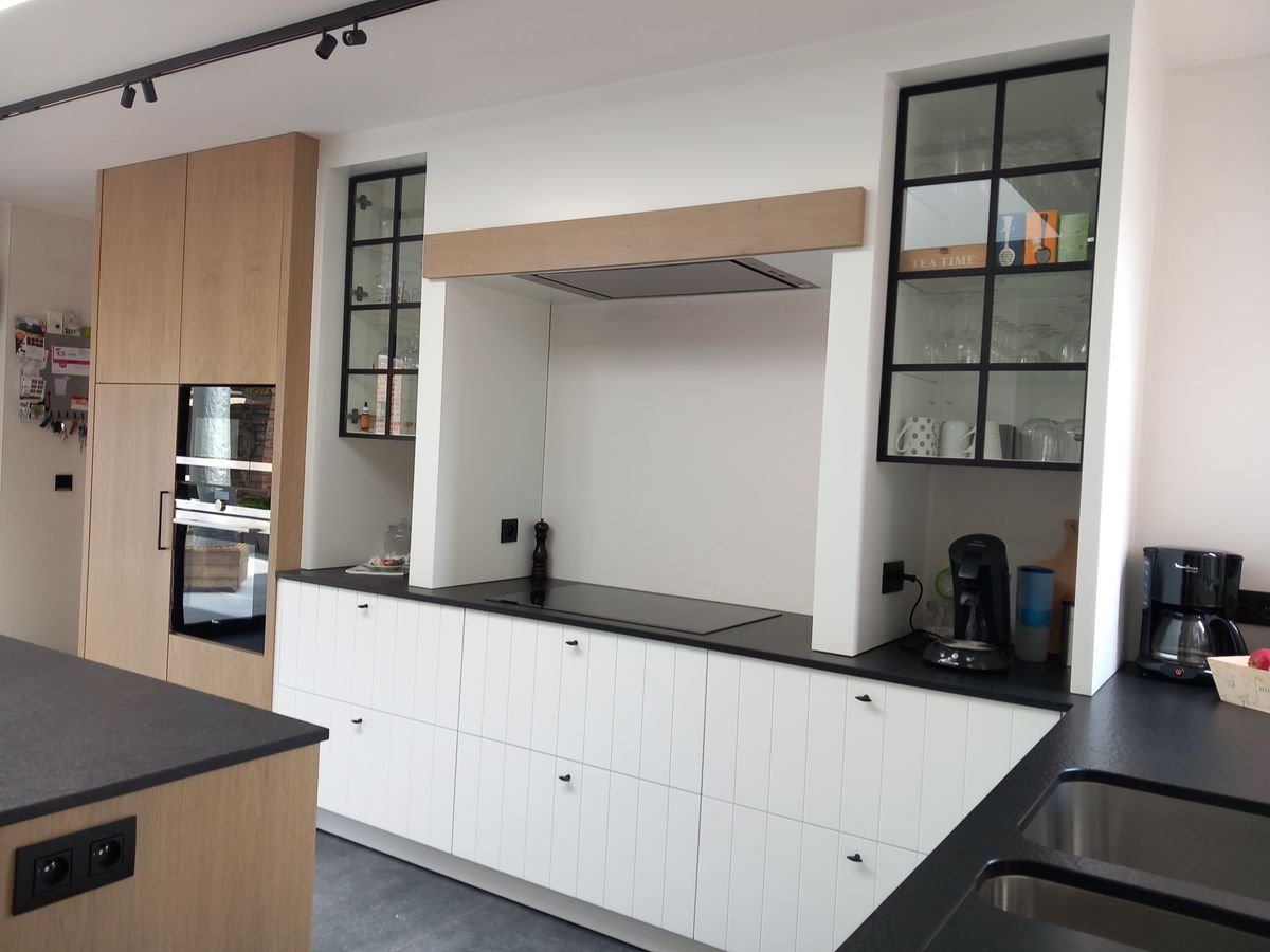 Keuken in Langemark met witgelakte kasten met groeven, eikenfineer kolomkasten, zwart werkblad en zwarte glasvitrinekasten naast de dampkap