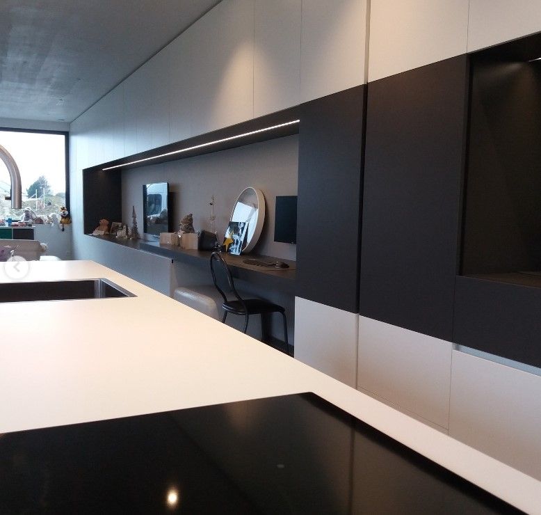 Lange zwart-witte kastenwand met verlichte nis en bureau, vooraan wit werkblad met zwarte kookplaat