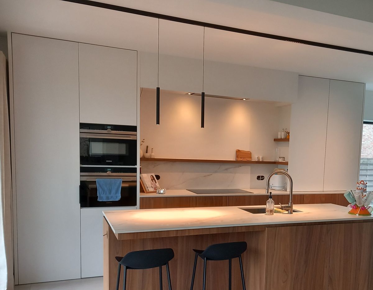 Witte hoge kastenwand met ingebouwde ovens, verlichte nis met houten legplanken en kookeiland in shinnoki