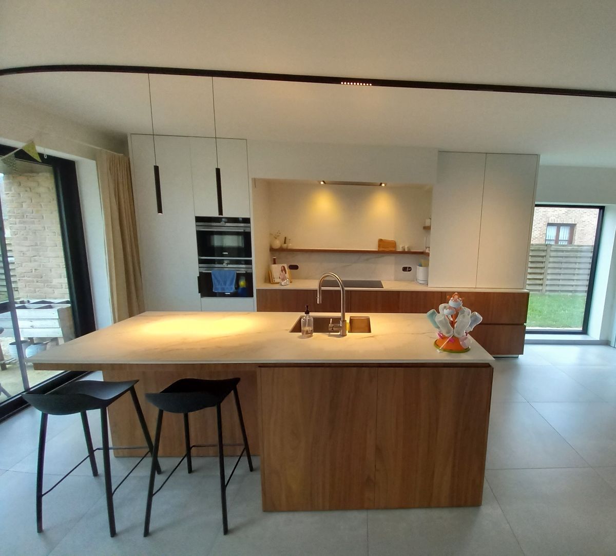 Keukeneiland in houtlook shinnoki met spoelbak, zwarte barkrukken en daarachter witte kasten met verlichte nis
