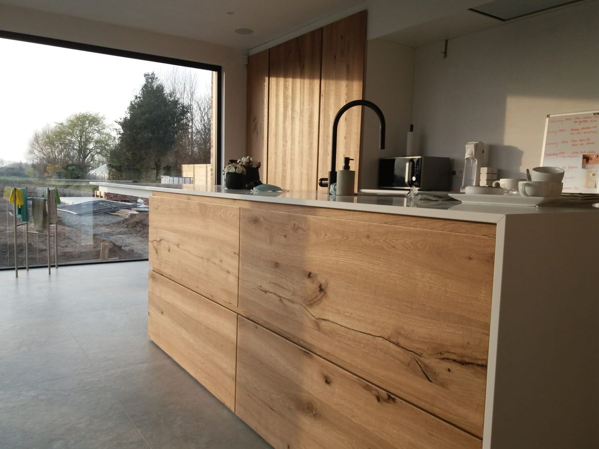 Keukeneiland in Vleteren met fronten in dik eikenfineer met knoesten, wit werkblad en zwarte kraan voor groot raam