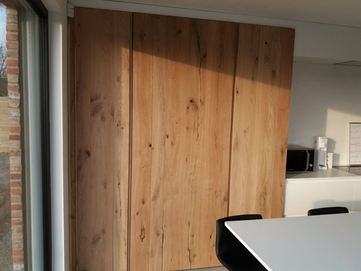 Hoge kastenwand in dik eikenfineer met grepenloze deuren, naast een raam en wit eiland