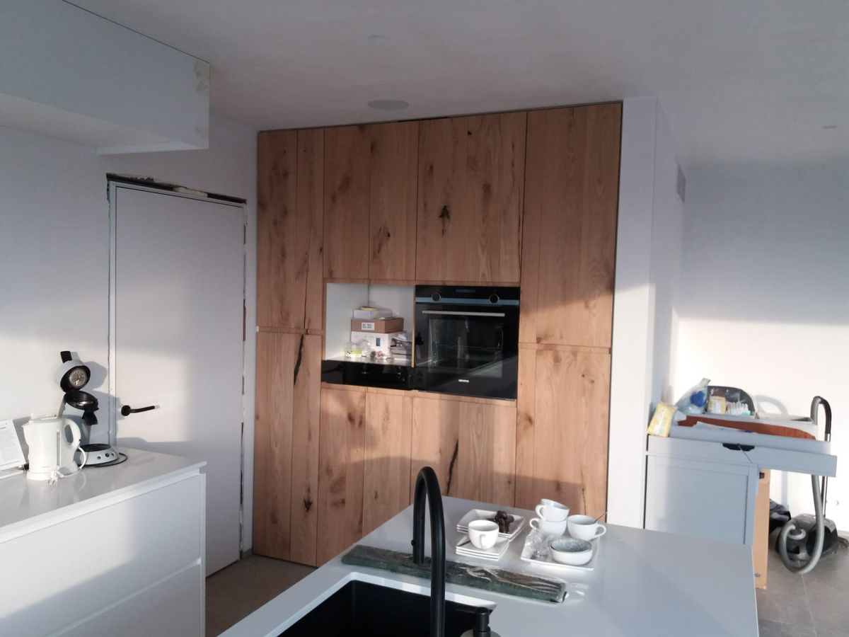 Kastenwand in dik eikenfineer met ingebouwde oven en open nis, gezien over het witte eiland