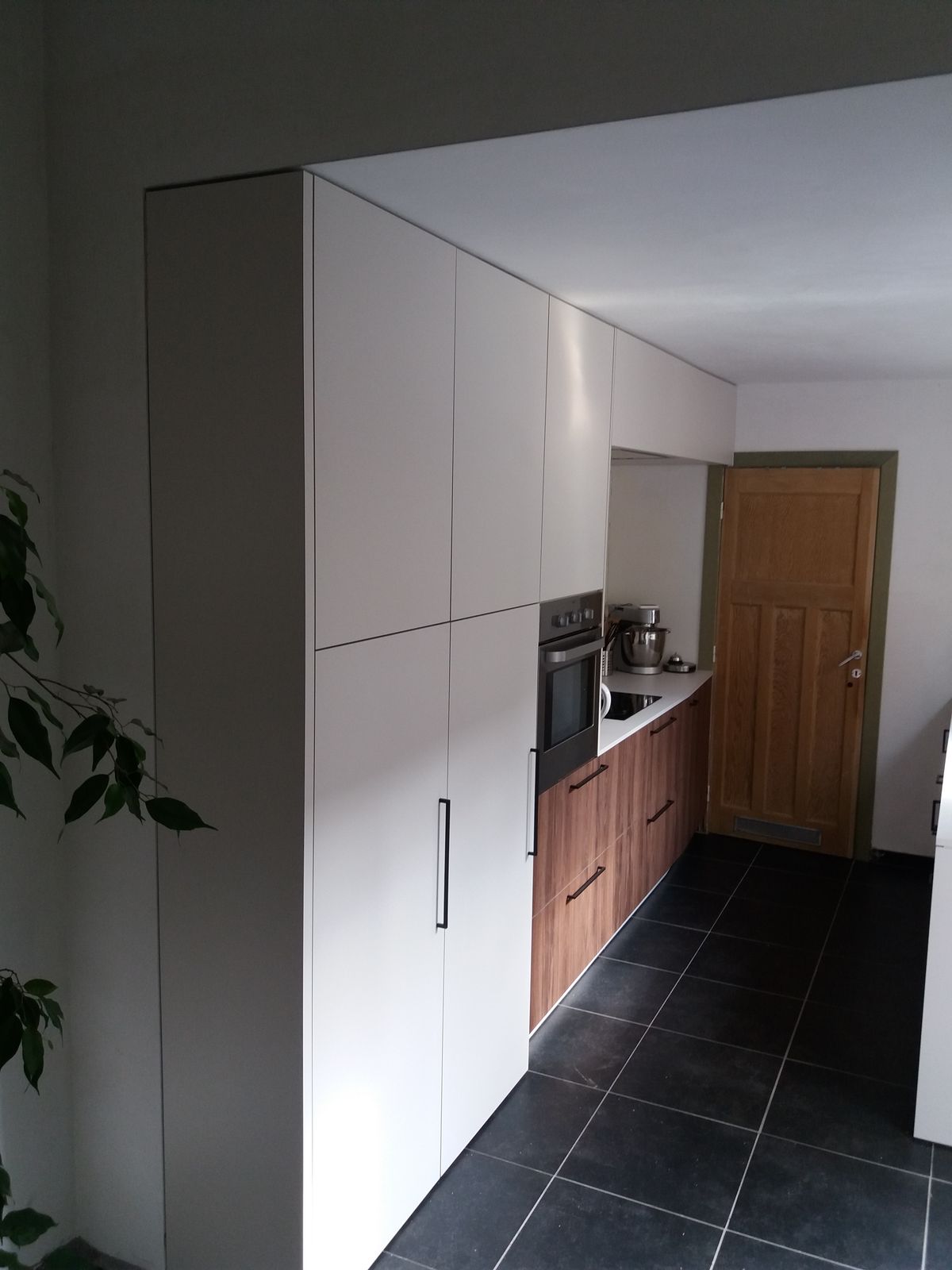 Smalle keuken in Ieper met witte hoge kasten in laminaat, houtkleurige onderkasten, ingebouwde oven en zwarte vloertegels