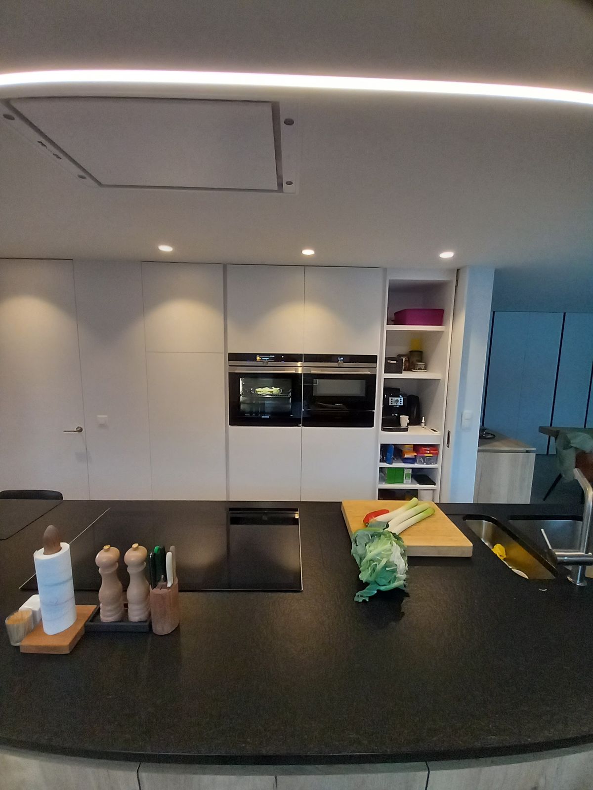 Zwart werkblad met kookplaat vooraan, witte hoge kasten met twee ovens en open rek erachter
