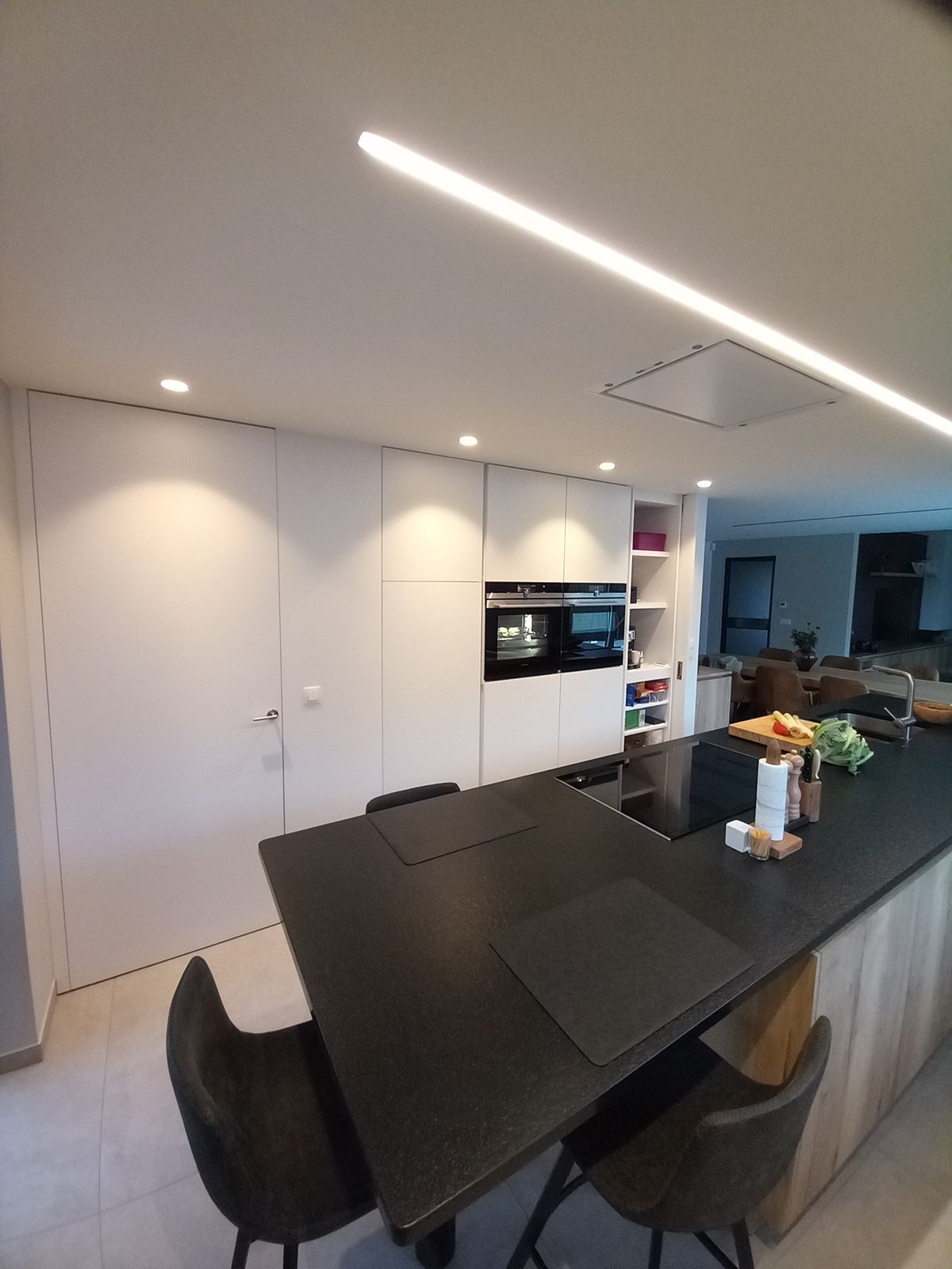 Witte greeploze kastenwand met ingebouwde ovens en zwart eiland met zitgedeelte onder lineaire ledverlichting