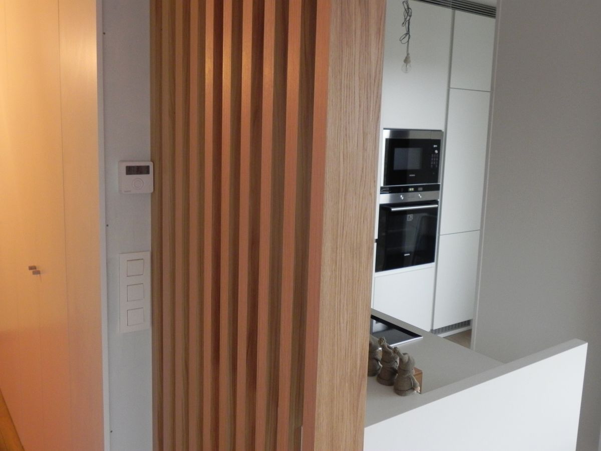 Verticale houten lattenafscheiding tussen inkomhal en witte keuken met ingebouwde oven en microgolfoven