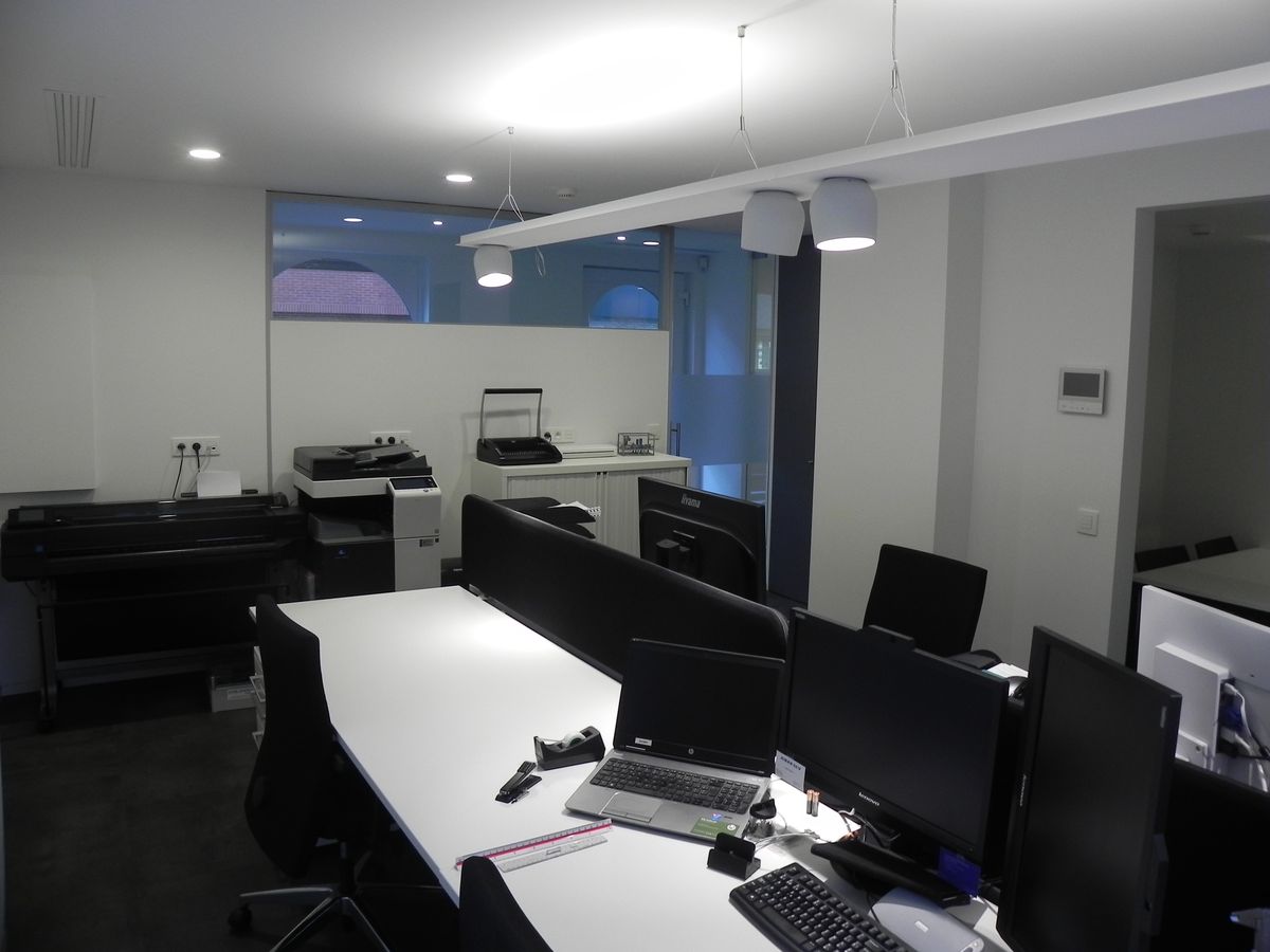 Kantoorruimte met witte bureaus, beeldschermen, printers en hoge ramen met bovenlicht