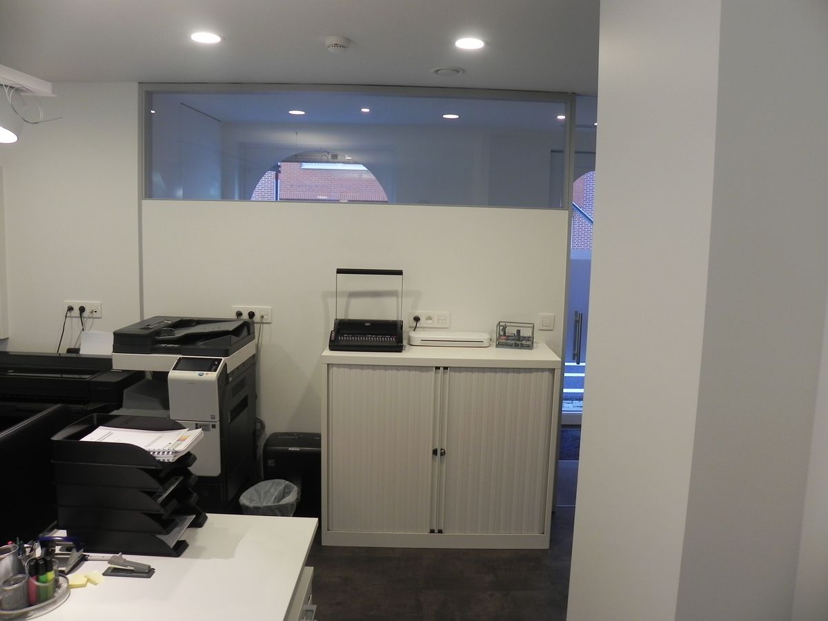 Werkplek met witte bureau en printers, witte kast met roldeur en raam met bovenlicht