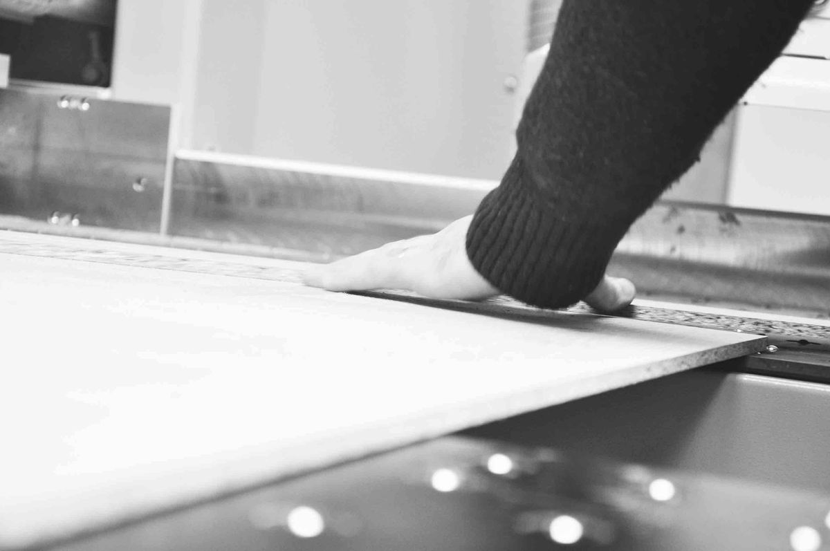 Zwart-witfoto van een hand die een plaat hout tegen de geleider van een zaagmachine in het atelier legt