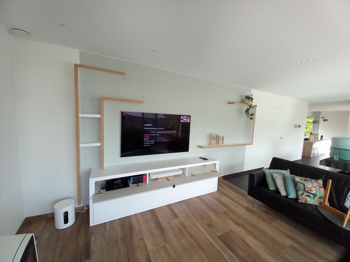 Wit tv-meubel met open lade, eronder audioapparatuur, en asymmetrische houten wandplanken rond de tv